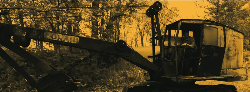A man at the controls of a weathered Lorain shovel on a gravel track below trees
