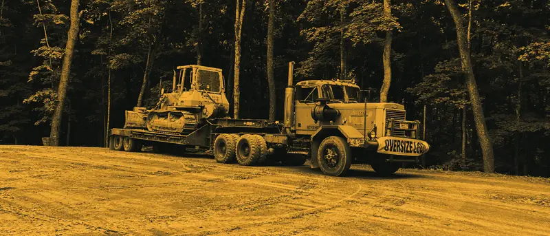 A crawler tractor chained to a low trailer behind an orange tractor unit with an oversize-load sign, on a dirt clearing below trees